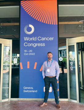 Kevin Linn standing in front of a large banner that says World Cancer Congress