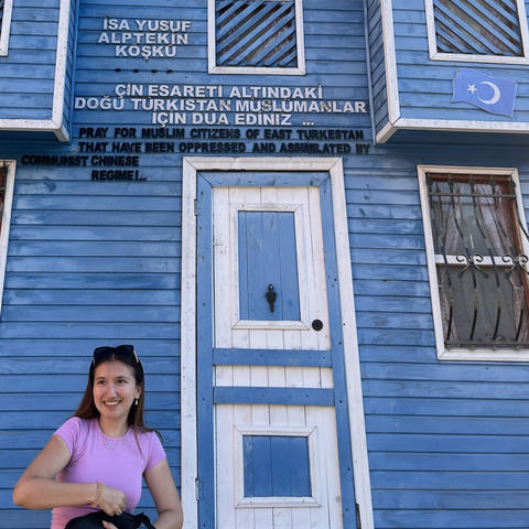 A smiling woman stands in front of a blue wooden building with signs in Turkish, English, and Chinese referencing oppression of Muslims in East Turkestan.