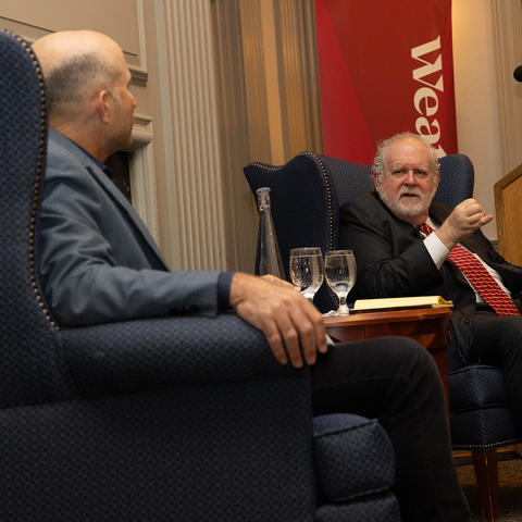 Two men in suits sit on blue armchairs, engaged in conversation near a podium, with water glasses and a bottle on the table between them.