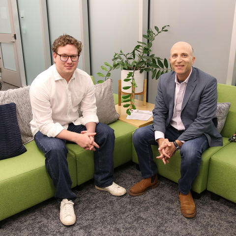 Two men in professional attire are seated on a green couch and smiling at the camera. 