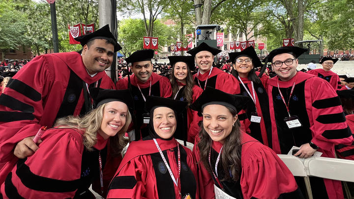 Nine graduate students in red and black caps and robes pose at graduation ceremony.