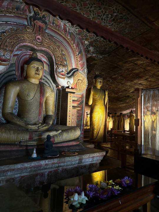 A dimly lit temple interior with large seated and standing Buddha statues, ornate carvings, painted ceiling, and flowers placed on a black altar.