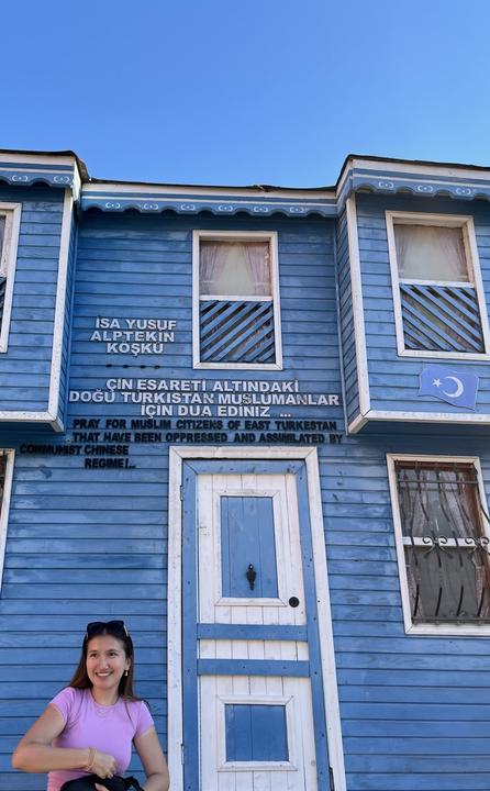 A smiling woman stands in front of a blue wooden building with signs in Turkish, English, and Chinese referencing oppression of Muslims in East Turkestan.