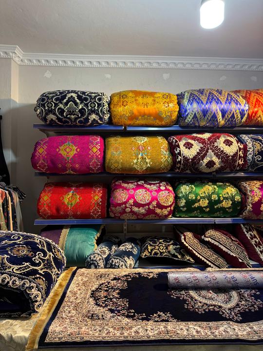Shelves display colorful, patterned textiles and folded blankets with intricate designs. A patterned rug lies on a table in front of the shelves.