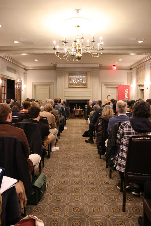 A group of people sit in rows facing a fireplace and two speakers at the front of a well-lit room with a chandelier and patterned carpet.