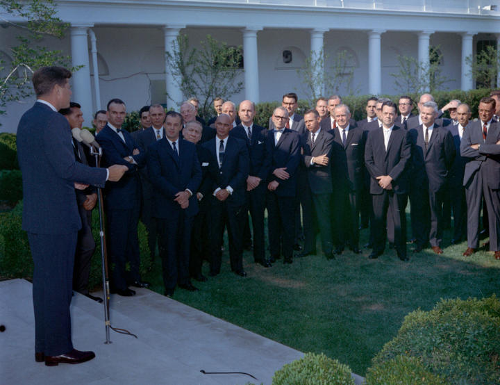 John F. Kennedy, Jr. speaks at a microphone to the Directors and Deputy Directors from Agency for International Development gathered outdoors in the White House Rose Garden.