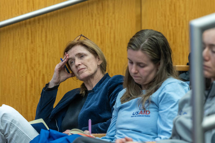 Two women sitting in an auditorium, one appearing to listen attentively with a pen in hand, the other looking down at a notebook, both wearing casual clothing.