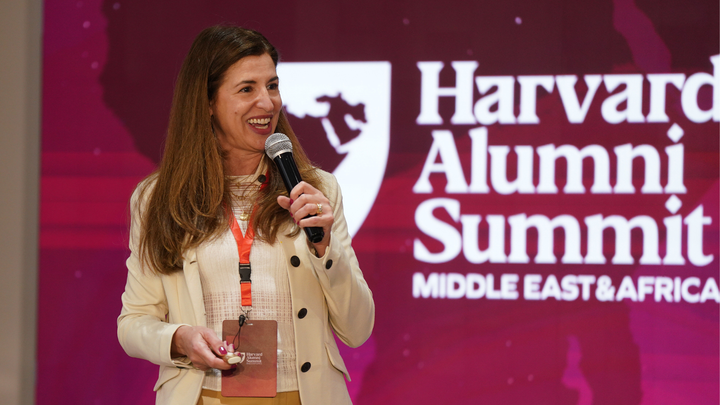 A woman holding a microphone speaks on stage at the Harvard Alumni Summit Middle East & Africa, with the event logo in the background.