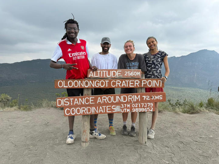 Four people stand smiling behind a sign at Oloolongot Crater Point, showing details about altitude and distance. Mountain landscape is visible in the background.