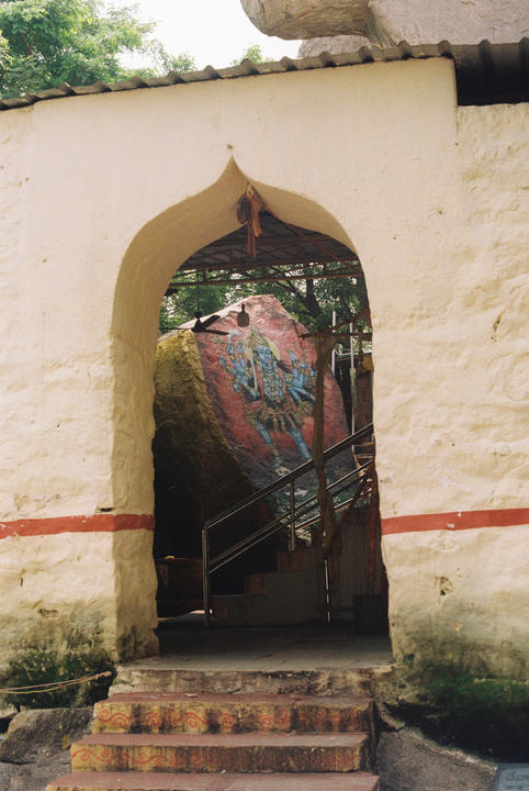 A stone structure with an arched entrance reveals a painted rock and stairs inside. Trees are visible in the background.