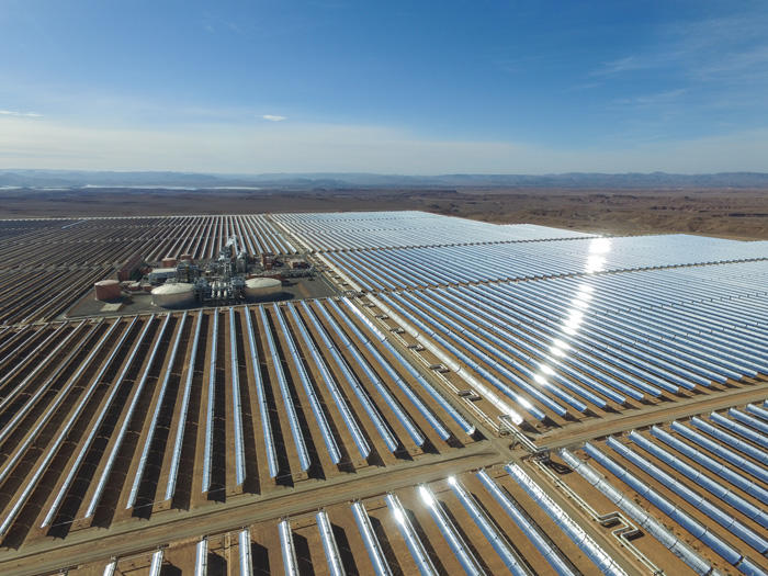 Aerial view of a vast solar power plant with rows of reflective solar panels in a desert landscape.