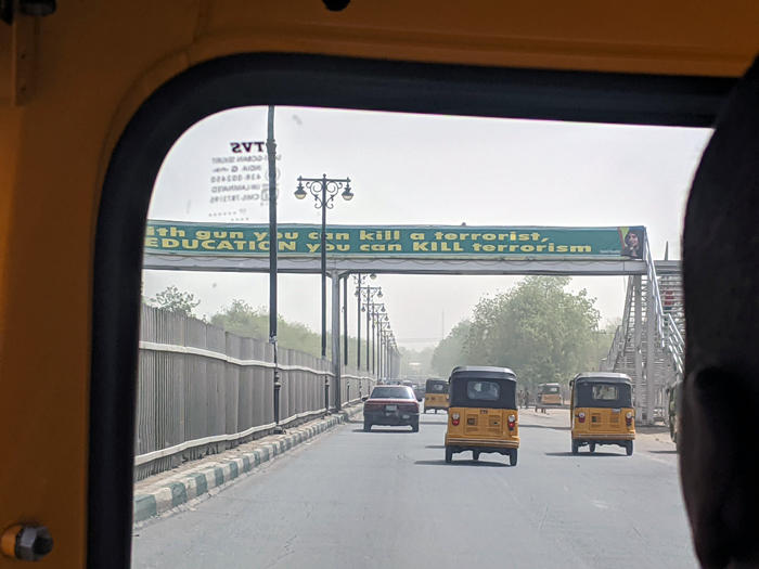 View from a rickshaw driving on a road in Maiduguri, passing under a banner quoting Malala