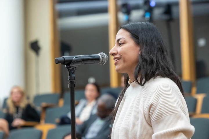 A woman with long dark hair speaks into a microphone in an auditorium setting with people in the background.