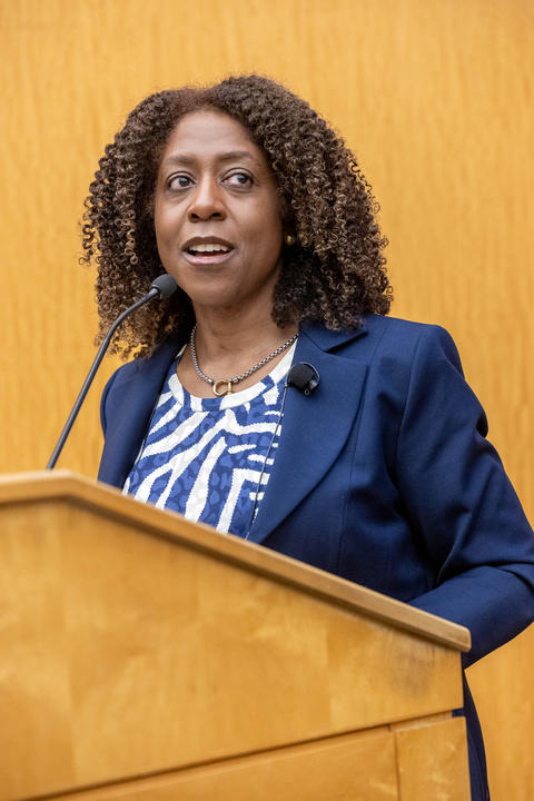 A woman with curly hair speaks at a podium, wearing a blue blazer and patterned blouse.