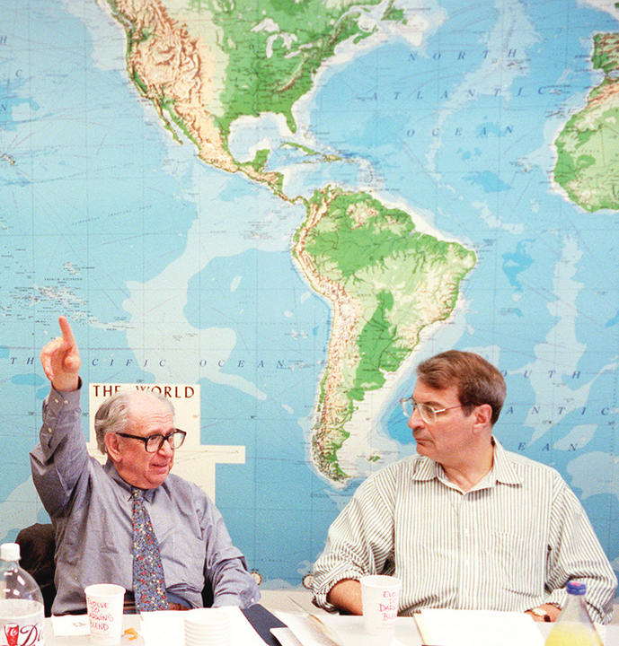 Herb Kelman and Peter Zimmerman sitting at table in front of a large world map