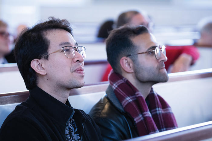 Two audience members, seated in church pews, listen to lecture.
