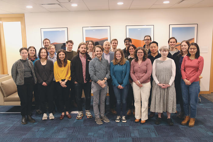 Group of 18 diverse graduate students smiling for a photo in a well-lit office space with artwork on the walls.