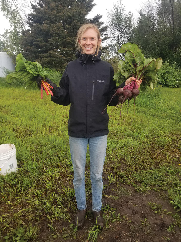 Mindy Jewell Price stands in a field of green holding carrots and beets in each hand.