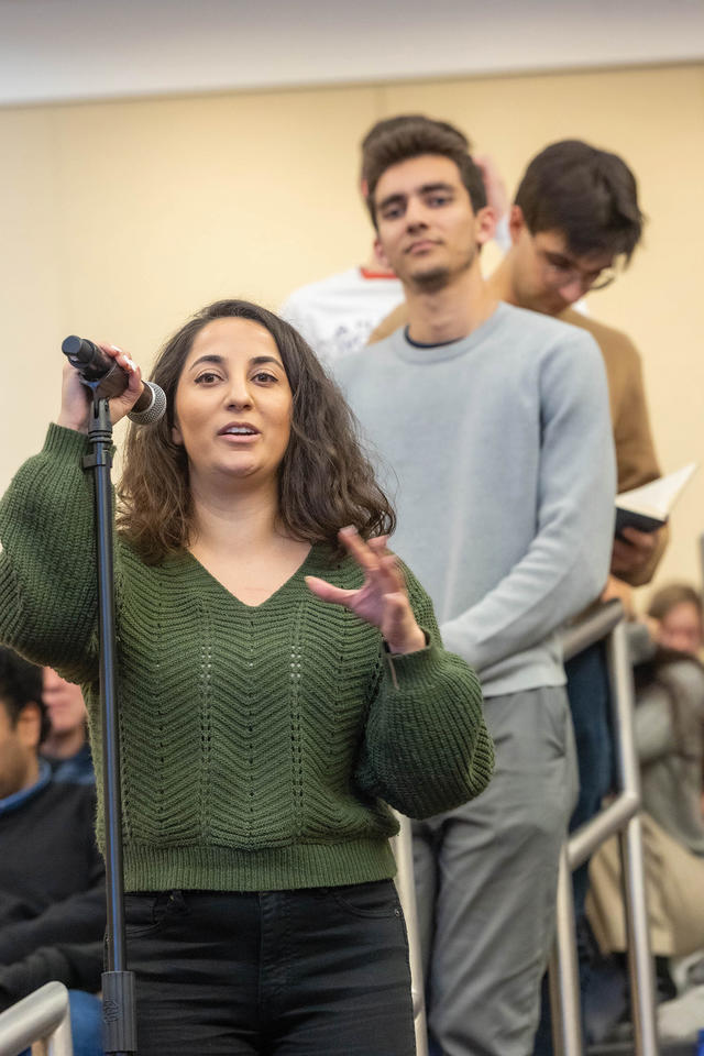 A woman in a green sweater stands at microphone to ask a question, with a line of people behind her.