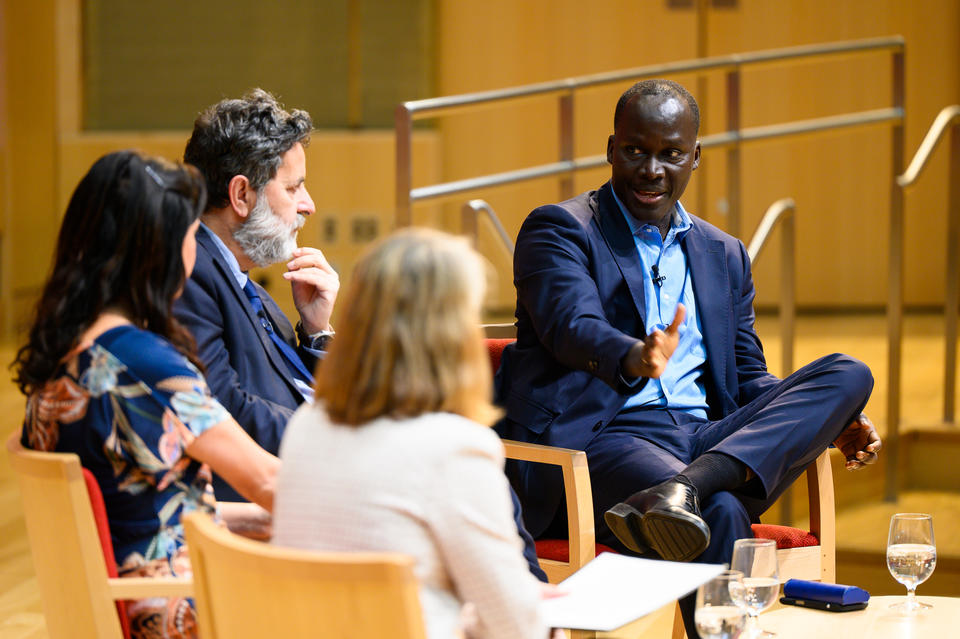 Four people in business attire are seated in a panel discussion on stage, with one man gesturing while speaking and the others listening attentively.