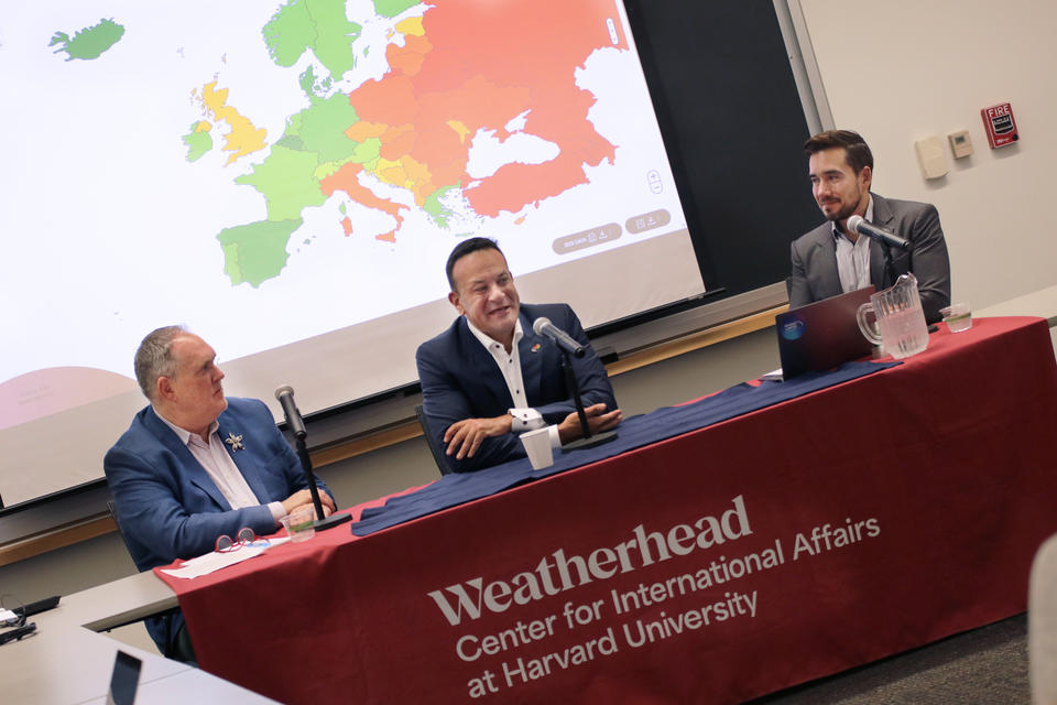 Three men sit at a table with microphones in front of a projected map of Europe, at a Weatherhead Center for International Affairs event at Harvard University.