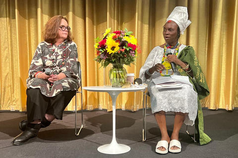 Two women with microphones sit on either side of a small white table with a large vase of flowers on it.