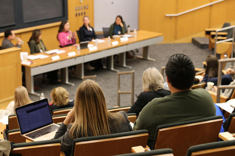 Looking across the audience toward the front of the room where five panelists are seated at the table.