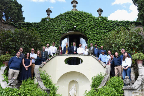 A couple dozen professionals stand outside on a grand stone staircase in front of an ivy-covered castle. 