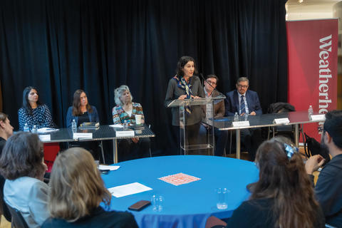 Several panelist sit at the front of a room with one speaking at a clear podium in the middle.
