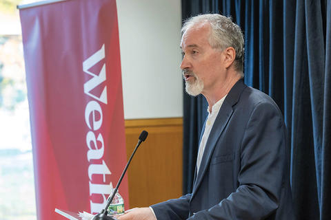 Man with gray hair and beard speaking at microphone on a podium.
