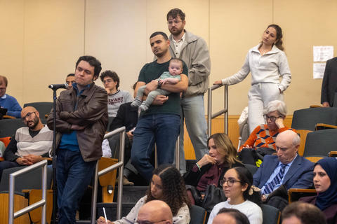 Several people stand in line in front of the microphone, among a crowded lecture hall.