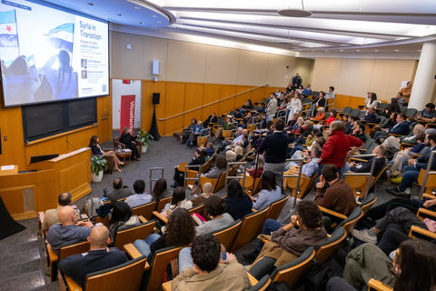 A crowded lecture hall with two women seated in conversation at the front.