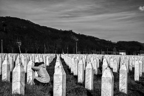 Woman kneels in front of a white gravestone in a large cemetery filled with identical gravestones. 