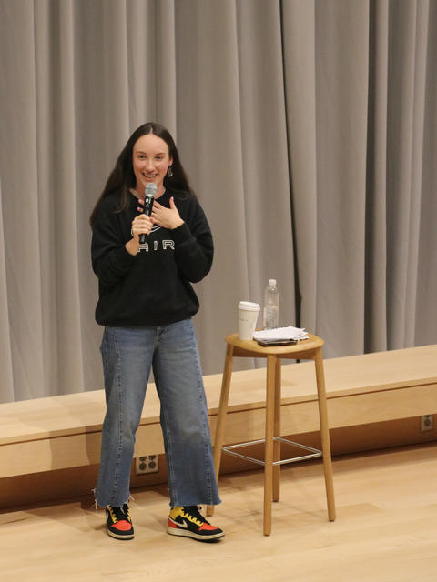 Woman holding microphone performs comedy routine on stage, standing next to a stool with water on it.
