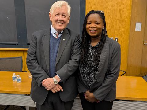 An older man in a suit and tie and a younger woman in a black outfit stand side by side in a classroom, smiling at the camera. There are desks, water bottles, and chalkboards behind them.