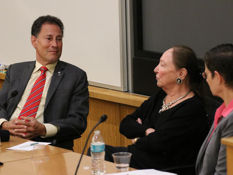 Three people in business attire sit at a conference table having a discussion, with water bottles and papers in front of them.