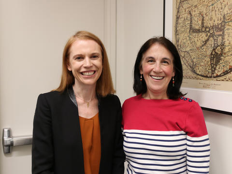 Two women standing indoors, smiling at the camera; one wears a black blazer and orange top, the other wears a red and white striped shirt, with a framed map on the wall behind them.