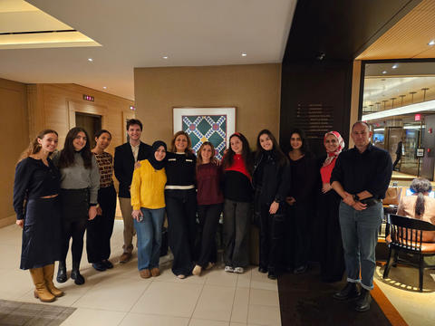 A group of thirteen people stands indoors in front of a framed artwork, smiling at the camera. The setting appears to be a hotel or restaurant lobby.
