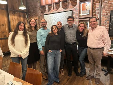 Eight adults standing together and smiling inside a restaurant with wooden and brick walls, art on the wall, and dining tables in the foreground.