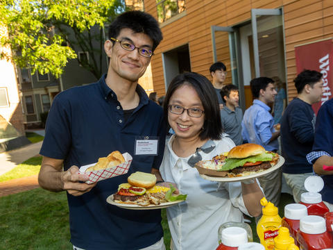 Two people stand outdoors holding plates of food at a gathering. Other attendees and food condiments are visible in the background.