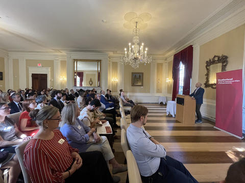 A speaker addresses an audience seated in a formal room with chandeliers and wooden floors; a red Weatherhead Center banner stands beside the podium.