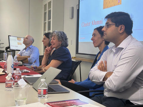 Five people sit at a conference table, listening attentively, with laptops and water bottles in front of them; a presentation slide reads "Cluster Alumni."