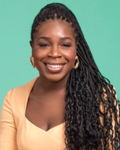 Woman with long brown braids is wearing a peach colored shirt and is smiling against a teal background.