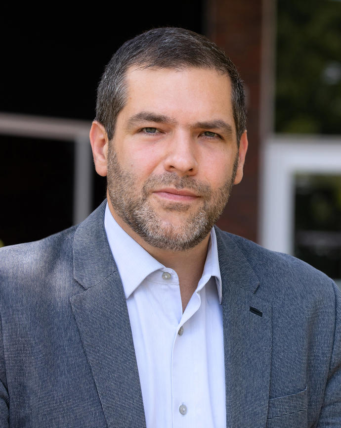 Man with short brown hair and sport coat standing in front of a building.