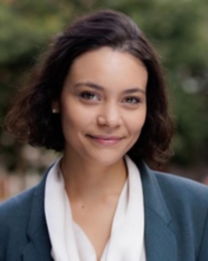 Woman with a brown bob wearing a gray blue suit is smiling in front of a tree.