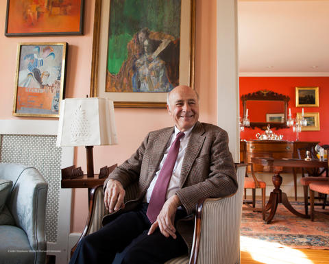 Joseph S. Nye, Jr. smiling and sitting in his living room chair with art on the walls and a small side table and lamp beside him.