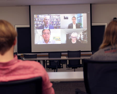 Two people sit in a classroom watching a video call with four participants projected on a large screen at the front of the room.