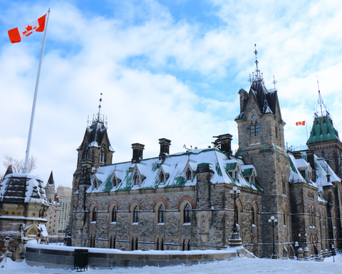 A photograph of Parliament Hill in Ottawa, Canada. There is snow on the rooftops and ground and a Canadian flag is flying to the left of the parliament building.